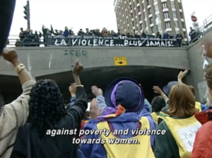 Still from the film "A Score for Women's Voices" featuring a multiracial group of women marching towards an underpass with their fists raised in solidarity. Above, them more women stand on a bridge cheering.