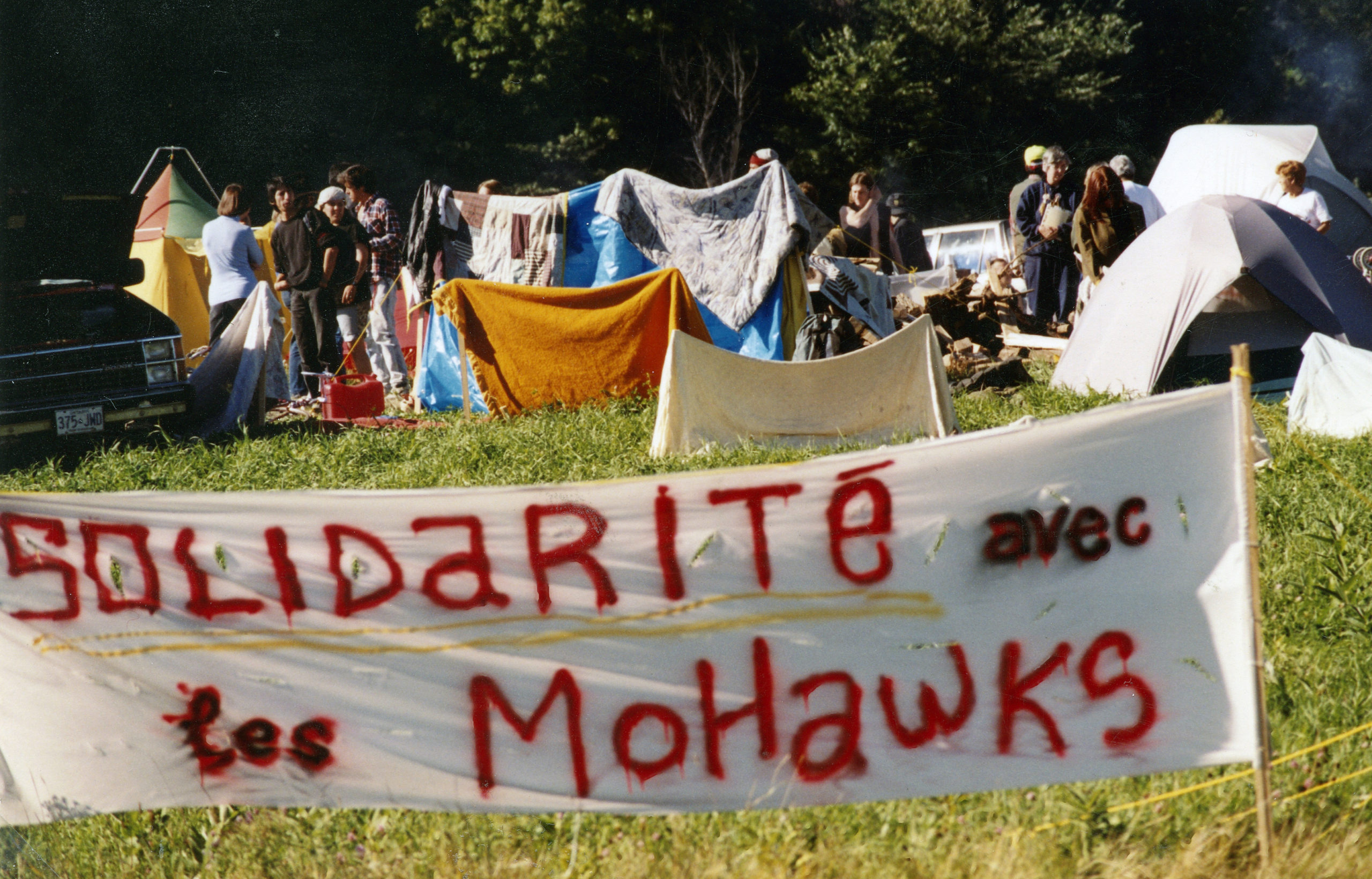 In the foreground of this photograph is a banner that says "Solidarité avec les Mohawks." In the background, people are standing by a cluster of multi-coloured tents on a grass field.