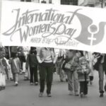 Film still of a black and white photo shown in 'IWD Vancouver Events/Exhibits 1985-1987' of a group of people marching. Two women are holding a banner that states "International Women's Day March 8 '86 - Vancouver"