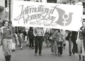 Film still of a black and white photo shown in 'IWD Vancouver Events/Exhibits 1985-1987' of a group of people marching. Two women are holding a banner that states "International Women's Day March 8 '86 - Vancouver"