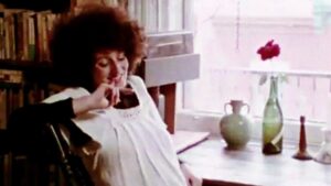 Film still from 'J’me marie, j’me marie pas' of a woman sitting at a desk turned to the camera smiling with her hand towards her chin. There are two flower vases on the desk with one holding a pink flower. The woman is also sitting in front of a book cart.