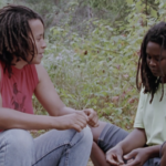 Film still from "Long Time Comin'" of Faith Nolan sitting on the left side talking to Grace Channer, sitting on the right side. Both are sitting in a forest.