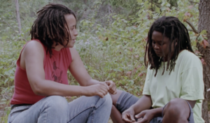 Film still from "Long Time Comin'" of Faith Nolan sitting on the left side talking to Grace Channer, sitting on the right side. Both are sitting in a forest.