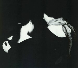 Black and white image of two women. The women on the right has her left hand resting on the left shoulder of the women on the right side.