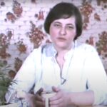 Film still of woman at the centre of the image sitting at a table holding a mug with both hands.