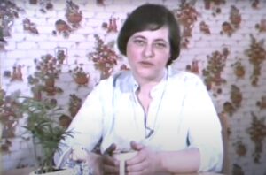 Film still of woman at the centre of the image sitting at a table holding a mug with both hands.