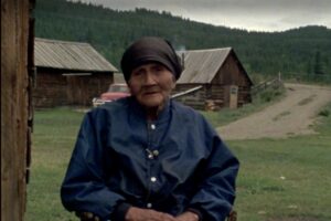 Film still of Augusta sitting with two log cabins behind her.