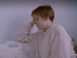 Film still of a woman sitting on a couch, holding her head with her right arm, staring down.