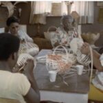 Film still of a group of women sitting around a table working on basket weaving.