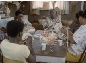 Film still of a group of women sitting around a table working on basket weaving.