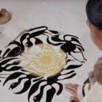 Film still of Inuit Artist, Kenojuak standing in front of her artwork printed on paper. Her hand rests on her art, depicting black birds positioned around a circle, with a yellow circular overlay in the middle of the circle.