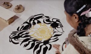 Film still of Inuit Artist, Kenojuak standing in front of her artwork printed on paper. Her hand rests on her art, depicting black birds positioned around a circle, with a yellow circular overlay in the middle of the circle.