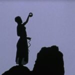 Film Still from Goddess Remembered of a silhouetted woman standing on a cliff holding a glass ball in one hand.