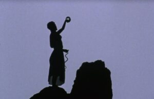 Film Still from Goddess Remembered of a silhouetted woman standing on a cliff holding a glass ball in one hand.