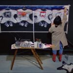 Film still from Hands of History of a woman standing in front of an art piece, panting the top right corner. The art piece contains images of wolves, clothing hangers and symbols of Indigenous clothing. There are also white painted handprints on the bottom portion of the art piece. There is also a small table with paint, paint brushes and other art supplies directly behind the woman.