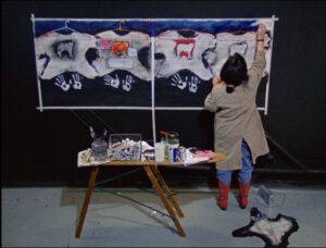 Film still from Hands of History of a woman standing in front of an art piece, panting the top right corner. The art piece contains images of wolves, clothing hangers and symbols of Indigenous clothing. There are also white painted handprints on the bottom portion of the art piece. There is also a small table with paint, paint brushes and other art supplies directly behind the woman.