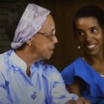 Film still from Home to Buxton of two Black women smiling at one another while sitting side by side.