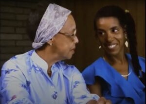 Film still from Home to Buxton of two Black women smiling at one another while sitting side by side.