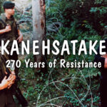 Film still from 'Kanehsatake: 270 Years of Resistance' with the title of the film written in white in the middle of the image. Two law enforcement men are positioned at the left of the image, while two other men are positioned on the right, with barbed wire in between them. All four men are standing in a forest.