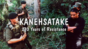 Film still from 'Kanehsatake: 270 Years of Resistance' with the title of the film written in white in the middle of the image. Two law enforcement men are positioned at the left of the image, while two other men are positioned on the right, with barbed wire in between them. All four men are standing in a forest.