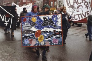 This photo taken at an IWD March in Toronto shows the Black Women's Collective and their banner, as well as Women Against the Arms Race.