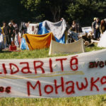 This photo is one of a number taken at the Oka Peace Camp set up in July 1990 in solidarity with the Mohawks of Kanehsatake who rose up in defense of their ancestral lands after the Oka Golf Club proposed an extension and the building of luxury condos over a Mohawk ancestral graveyard in the sacred wooded area known as “The Pines”. This land had never been ceded. In early July, after the Mohawks refused to end their non-violent occupation of the area or to take down the barricade, the Sûreté du Québec (Quebec police) moved in resulting in a violent confrontation. Later, the Canadian army was called in. Mohawk women, including Ellen Gabriel, played a central role in the uprising, which lasted for 78 intense days (July 11 – September 26, 1990). In the end, the golf course was not extended, and the condos were not built. But the larger issues of land sovereignty have never been resolved.