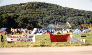 This photo is one of a number taken at the Oka Peace Camp set up in July 1990 in solidarity with the Mohawks of Kanehsatake who rose up in defense of their ancestral lands after the Oka Golf Club proposed an extension and the building of luxury condos over a Mohawk ancestral graveyard in the sacred wooded area known as “The Pines”. This land had never been ceded. In early July, after the Mohawks refused to end their non-violent occupation of the area or to take down the barricade, the Sûreté du Québec (Quebec police) moved in resulting in a violent confrontation. Later, the Canadian army was called in. Mohawk women, including Ellen Gabriel, played a central role in the uprising, which lasted for 78 intense days (July 11 – September 26, 1990). In the end, the golf course was not extended, and the condos were not built. But the larger issues of land sovereignty have never been resolved.