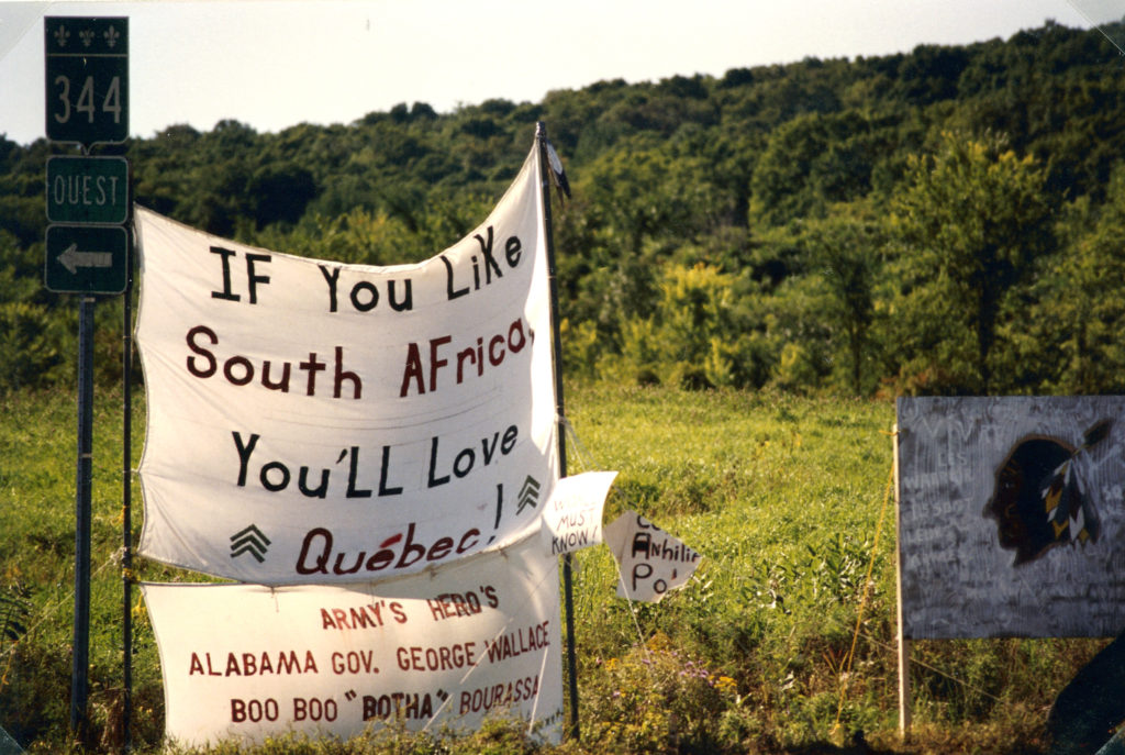 This photo is one of a number taken at the Oka Peace Camp set up in July 1990 in solidarity with the Mohawks of Kanehsatake who rose up in defense of their ancestral lands after the Oka Golf Club proposed an extension and the building of luxury condos over a Mohawk ancestral graveyard in the sacred wooded area known as “The Pines”. This land had never been ceded. In early July, after the Mohawks refused to end their non-violent occupation of the area or to take down the barricade, the Sûreté du Québec (Quebec police) moved in resulting in a violent confrontation. Later, the Canadian army was called in. Mohawk women, including Ellen Gabriel, played a central role in the uprising, which lasted for 78 intense days (July 11 – September 26, 1990). In the end, the golf course was not extended, and the condos were not built. But the larger issues of land sovereignty have never been resolved.