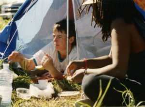 This photo is one of a number taken at the Oka Peace Camp set up in July 1990 in solidarity with the Mohawks of Kanehsatake who rose up in defense of their ancestral lands after the Oka Golf Club proposed an extension and the building of luxury condos over a Mohawk ancestral graveyard in the sacred wooded area known as “The Pines”. This land had never been ceded. In early July, after the Mohawks refused to end their non-violent occupation of the area or to take down the barricade, the Sûreté du Québec (Quebec police) moved in resulting in a violent confrontation. Later, the Canadian army was called in. Mohawk women, including Ellen Gabriel, played a central role in the uprising, which lasted for 78 intense days (July 11 – September 26, 1990). In the end, the golf course was not extended, and the condos were not built. But the larger issues of land sovereignty have never been resolved.