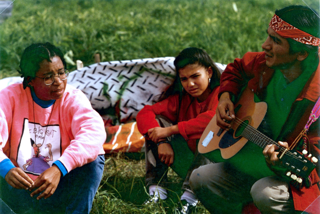 This photo is one of a number taken at the Oka Peace Camp set up in July 1990 in solidarity with the Mohawks of Kanehsatake who rose up in defense of their ancestral lands after the Oka Golf Club proposed an extension and the building of luxury condos over a Mohawk ancestral graveyard in the sacred wooded area known as “The Pines”. This land had never been ceded. In early July, after the Mohawks refused to end their non-violent occupation of the area or to take down the barricade, the Sûreté du Québec (Quebec police) moved in resulting in a violent confrontation. Later, the Canadian army was called in. Mohawk women, including Ellen Gabriel, played a central role in the uprising, which lasted for 78 intense days (July 11 – September 26, 1990). In the end, the golf course was not extended, and the condos were not built. But the larger issues of land sovereignty have never been resolved.