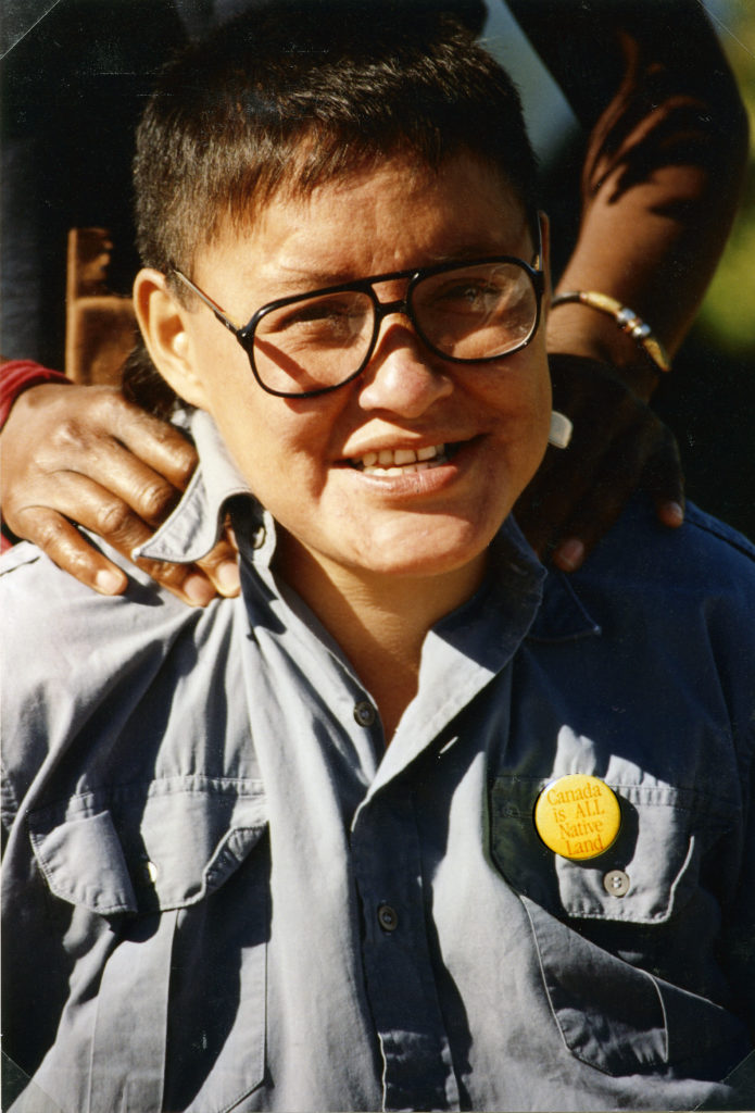 This photo is one of a number taken at the Oka Peace Camp set up in July 1990 in solidarity with the Mohawks of Kanehsatake who rose up in defense of their ancestral lands after the Oka Golf Club proposed an extension and the building of luxury condos over a Mohawk ancestral graveyard in the sacred wooded area known as “The Pines”. This land had never been ceded. In early July, after the Mohawks refused to end their non-violent occupation of the area or to take down the barricade, the Sûreté du Québec (Quebec police) moved in resulting in a violent confrontation. Later, the Canadian army was called in. Mohawk women, including Ellen Gabriel, played a central role in the uprising, which lasted for 78 intense days (July 11 – September 26, 1990). In the end, the golf course was not extended, and the condos were not built. But the larger issues of land sovereignty have never been resolved.