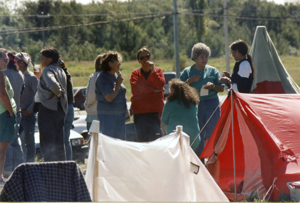 This photo is one of a number taken at the Oka Peace Camp set up in July 1990 in solidarity with the Mohawks of Kanehsatake who rose up in defense of their ancestral lands after the Oka Golf Club proposed an extension and the building of luxury condos over a Mohawk ancestral graveyard in the sacred wooded area known as “The Pines”. This land had never been ceded. In early July, after the Mohawks refused to end their non-violent occupation of the area or to take down the barricade, the Sûreté du Québec (Quebec police) moved in resulting in a violent confrontation. Later, the Canadian army was called in. Mohawk women, including Ellen Gabriel, played a central role in the uprising, which lasted for 78 intense days (July 11 – September 26, 1990). In the end, the golf course was not extended, and the condos were not built. But the larger issues of land sovereignty have never been resolved.