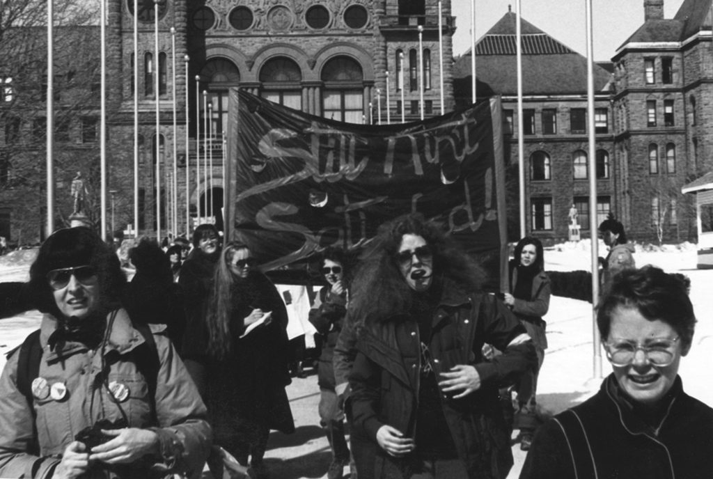 On an International Women's Day March in front of Queen's Park, marchers carry the banner "Still Ain't Satisfied".