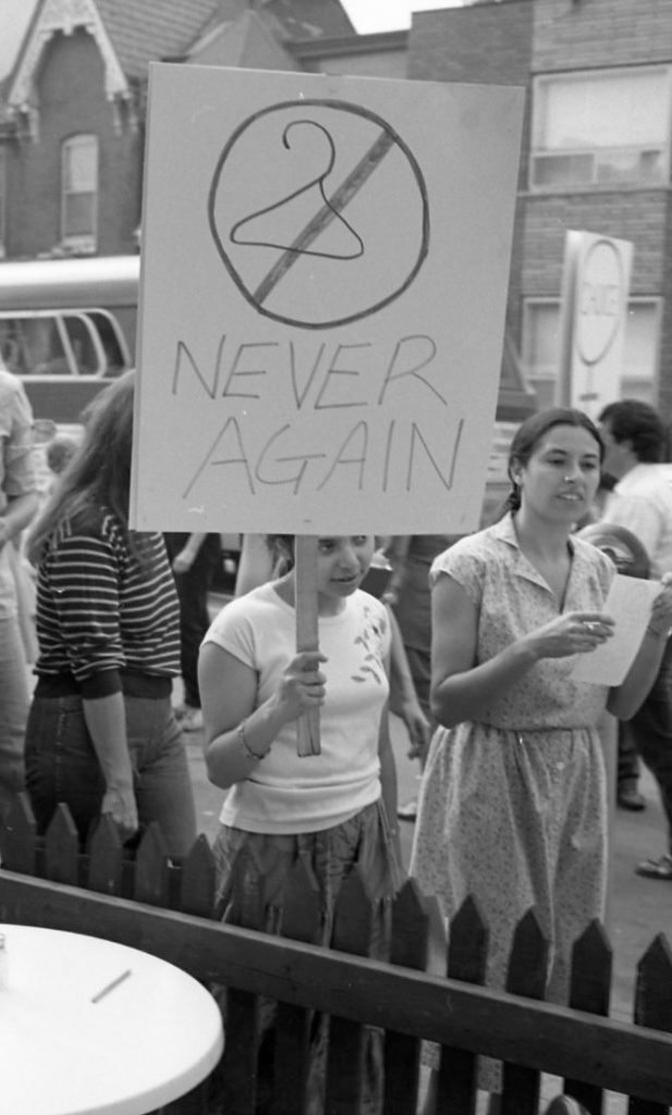 Placard at 1983 support rally outside the Morgentaler Clinic in Toronto shows a coat hanger and reads, "Never Again".