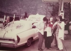 This photo taken was during a gas stop near Hope, British Columbia during Abortion Caravan's trip across Canada. Betsy Wood is on the left side of her convertible with an "On to Ottawa" sign fastened on the front. On the right side, from left to right: Dodie Weppler, Ellen Woodsworth, Gwen Hauser, and Bonnie Beckwoman.