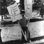 In this photograph, a young person is holding two placards, one saying "Lesbians United Cannot Be Defeated" and the other saying "My Parents Have a Gay Kid".