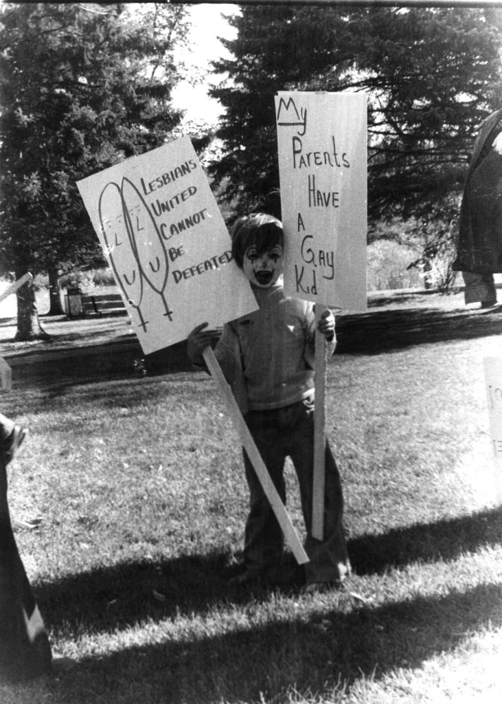 In this photograph, a young person is holding two placards, one saying "Lesbians United Cannot Be Defeated" and the other saying "My Parents Have a Gay Kid".