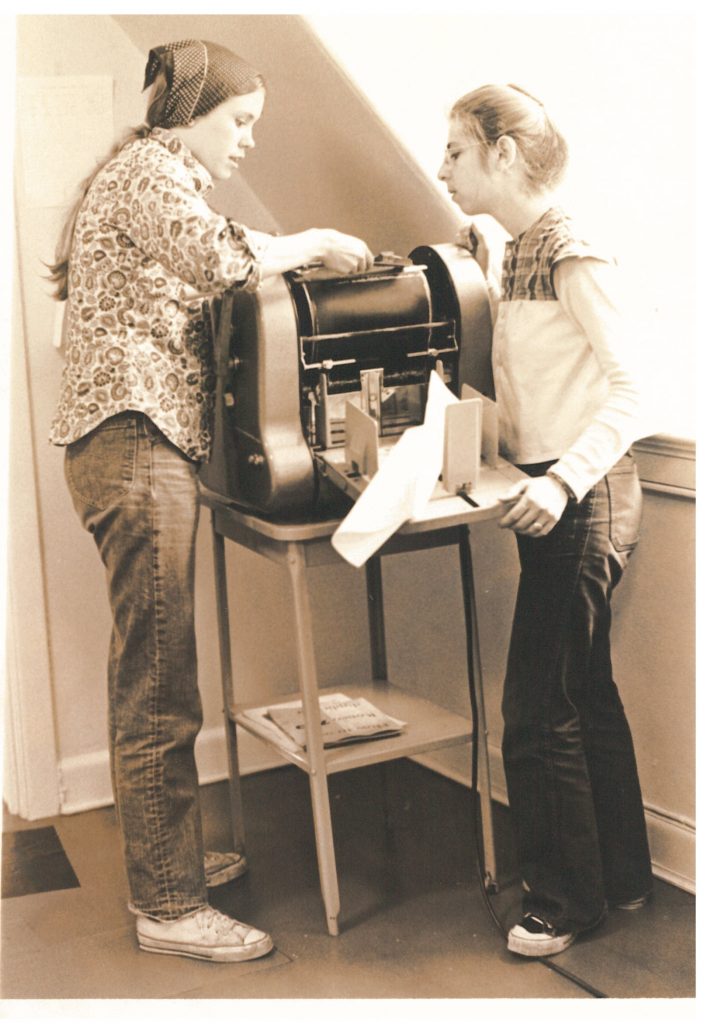 Cathy Carroll, now Billie Carroll, and Kathy Jackman working on the Gestetner at the Women's Press office on Bloor Street in Toronto. Photo taken between 1972 and 1976.