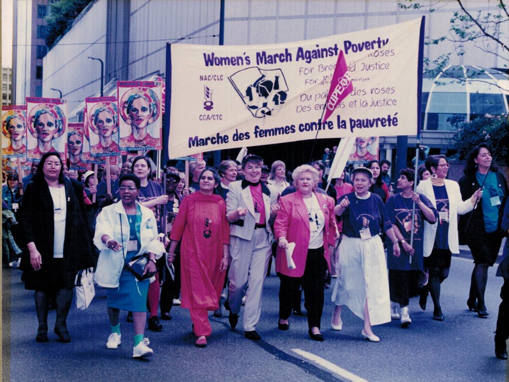 Photo taken at Women's March Against Poverty – For Bread and Roses, For Jobs and Justice.” The cross-country action was organized by the Canadian Labour Congress and the National Action Committee on the Status of Women in 1996. Right to left: Frances Lankin, Judy Darcy, Mary Ismail (holding banner), Nancy Riche, Louisette Hinton, Angie Schera, Sunera Thobani, Sue Carter (holding banner at left), June Veecock. Alexa McDonough, leader of the federal NDP is also visible between Sunera Thobani and Angie Schera.